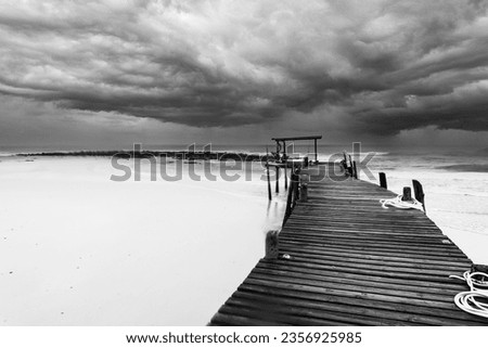 Black and white picture of Long wooden pier (bridge) extents from beach to turquoise wave sea. Deep Blue cloudy sky after the big storm.
