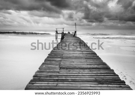 Black and white picture of Long wooden pier (bridge) extents from beach to turquoise wave sea. Deep Blue cloudy sky after the big storm.