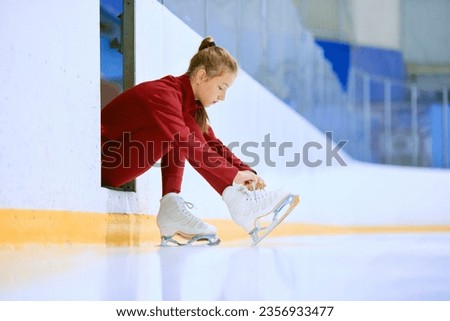 Beautiful girl in red sportswear preparing for training session on ice rink arena, figure skater tying laces on skates. Concept of professional sport, competition, sport school, health, hobby, ad Royalty-Free Stock Photo #2356933477