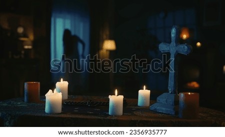 Shot capturing a table with magical tools on it: candles, cross and beads. On the background there is a blurry female silhouette, ghost moving weirdly in front of the window.
