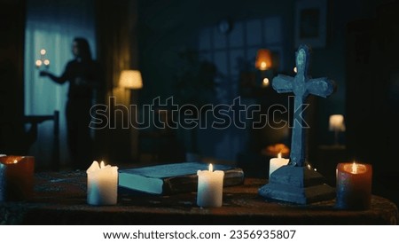 Shot capturing a table with magical tools on it: candles, cross and beads. A priest with a candlestick stands by the window. Place for occult rites and rituals. Royalty-Free Stock Photo #2356935807
