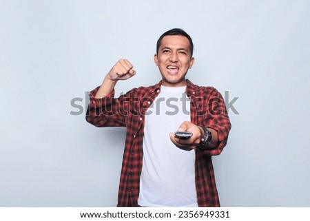 Asian young man wearing flannel shirt holding remote control and watching TV on white background.