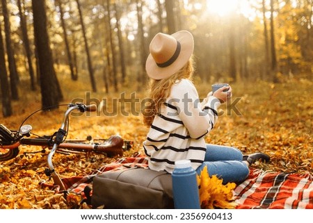 Beautiful curly woman in a hat sits on a red mat with a thermos in the autumn forest. Female enjoys nature at mini picnic. Concept of relaxation, nature.