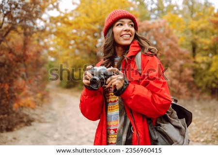 Happy female tourist with a camera and a backpack in a stylish sweater on a walk in the autumn forest. A woman takes pictures and enjoys the beautiful nature.