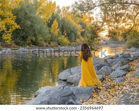 A woman in long yellow dress enjoy a scenery in Autumn or fall season color of leafs change on trees in chilly weather in a park. Sunlight and yellow trees reflected on peaceful river in a morning