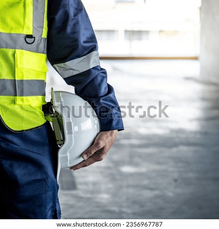 Safety workwear concept. Male hand holding white safety helmet or hard hat. Construction worker man in protective suit and reflective green vest standing with building concrete floor in the background Royalty-Free Stock Photo #2356967787