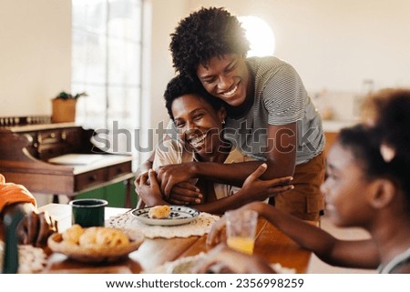 Brazilian family having breakfast, with mom and son sharing hugs at their kitchen table. Happy black family enjoying quality time together, with fresh cheese bread rolls served on the table. Royalty-Free Stock Photo #2356998259