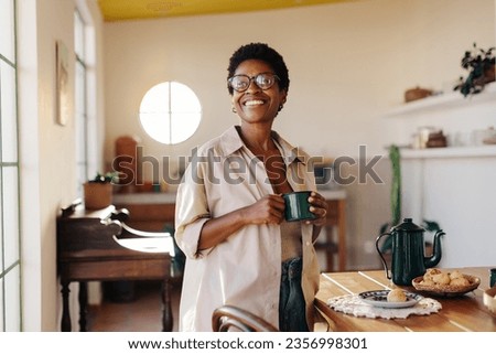 Brazilian woman holding a coffee cup and looking away thoughtfully in her home kitchen. home. Mature black woman standing by a breakfast table filled with freshly baked cheese bread rolls. Royalty-Free Stock Photo #2356998301