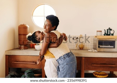 Happy young girl sharing a loving hug with her mom in a Brazilian kitchen. Mother and daughter filling the room with laughter as they enjoy quality time together, creating memories in their home. Royalty-Free Stock Photo #2356998391