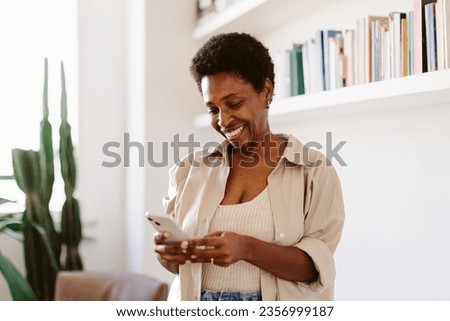 Happy, mature black woman stands in her living room next to a bookshelf, browsing her mobile phone. Woman reading a text message with a smile. at home. Royalty-Free Stock Photo #2356999187