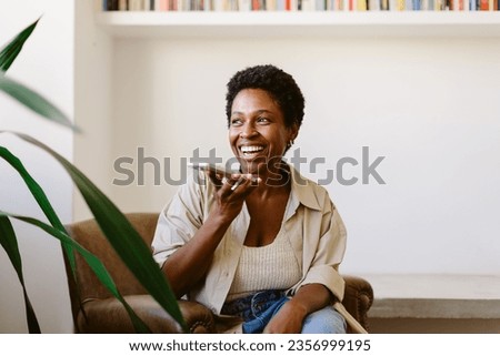 Happy Brazilian woman sits on a couch, speaking on her smartphone. Smiling and relaxed, she holds the phone and looks away, fully engaged in a phone call from her living room. Royalty-Free Stock Photo #2356999195