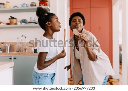 Black mother and her daughter have a fun sing-along at home, using wooden spoons as microphones. Mom and daughter enjoying themselves, creating lasting memories in the kitchen.