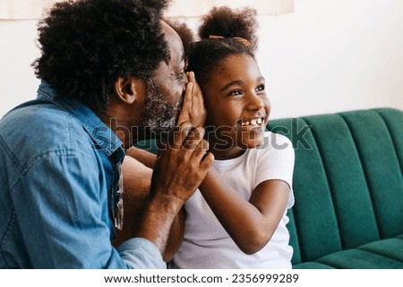 Dad whispering to his young daughter, creating a playful atmosphere and a genuine moment of father-daughter bonding at home. Parent and child spending some quality on a couch. Royalty-Free Stock Photo #2356999289