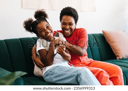Mother tickling her daughter who is ready for school with her school bag. Happy mom and daughter laughing and playing together in their cozy living room, creating a happy morning routine. Royalty-Free Stock Photo #2356999313