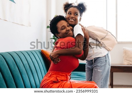 Joyful mom and daughter share a hug in their living room before the little girl heads off to school. The afro-haired girl is all set with her school bag, ready for a new day of learning. Royalty-Free Stock Photo #2356999331
