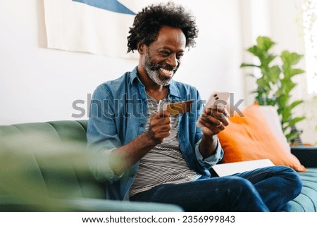 Mature man with afro hair happily using a mobile banking app on his smartphone to make a payment. Black man enjoying online shopping with his credit card at home. Royalty-Free Stock Photo #2356999843