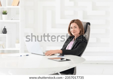 Confident and successful business lady sits at the desk and works on laptop in her modern office	