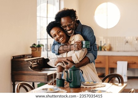Black Brazilian man holding his wife in a tight hug at the breakfast table. Mature couple standing together in their home kitchen, smiling, laughing and sharing a loving relationship. Royalty-Free Stock Photo #2357035415
