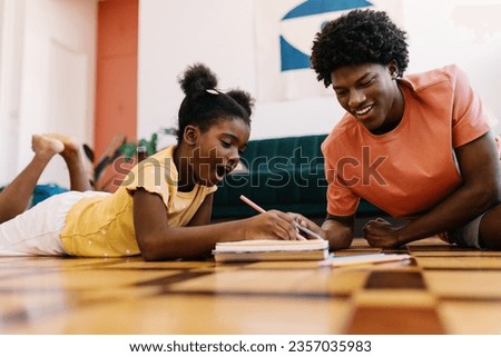 Big brother with afro hair happily assists his younger sister with her schoolwork, including drawing with colour pencils. Happy siblings sharing a genuine moment of learning and bonding at home. Royalty-Free Stock Photo #2357035983