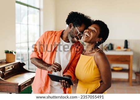Black mature couple with afro hair laughing together at home, browsing social media on a tablet device. Afro-haired husband and wife having fun while spending time indoors on a leisurely day. Royalty-Free Stock Photo #2357055491