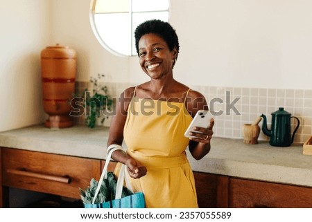Smiling Brazilian woman stands in her kitchen, holding a bag of fresh vegetables. She confidently poses with her mobile phone, capturing the essence of her cheerful cooking and happy lifestyle. Royalty-Free Stock Photo #2357055589
