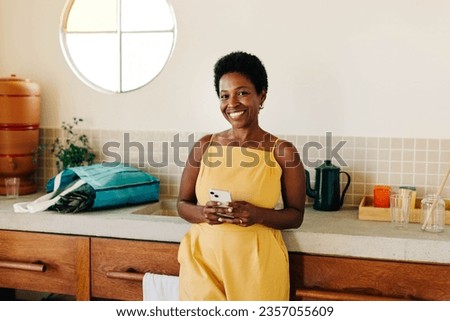 A happy Brazilian woman in a kitchen, using her mobile phone with a cheerful smile. Royalty-Free Stock Photo #2357055609