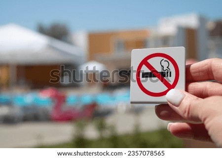 No smoking ban sign on the background of the pool, umbrellas and flamingos. A woman's hand holds a no smoking sign. Attention, do not smoke.