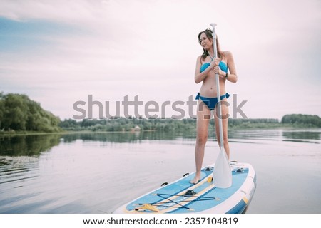 A young woman with an open swimsuit swims on a SUP board on a picturesque lake. SUP - Substitute the paddle. A beautiful, slender girl is engaged in sap surfing on the calm water 