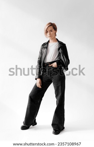 Full body of young woman with short blonde hair shows off her confident and stylish attitude in a black leather jacket and wide legged jeans, looking at camera against white background