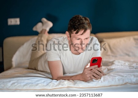 Man reading information in smartphone lying in bed at home in modern bedroom in evening. Interested concentrated male searching info, surfing online, watching content, scrolling social media in phone. Royalty-Free Stock Photo #2357113687