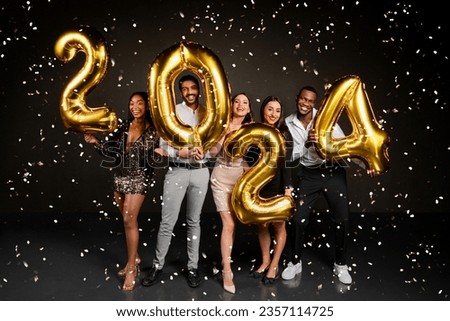 Excited beautiful multicultural friends men and women in nice outfits celebrating New Year 2024 together, holding golden number balloons and posing over black background, enjoying falling confetti Royalty-Free Stock Photo #2357114725