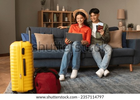 Happy asian family couple using laptop holding passports with tickets, booking flight and hotel online at home, tourists with packed suitcase planning vacation, browsing trip destination Royalty-Free Stock Photo #2357114797