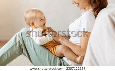 Happy laughing adorable blonde little baby playing in mom hands, motherhood concept. Unrecognizable mother bonding with her toddler son, sitting on floor in bedroom, holding her child on lap