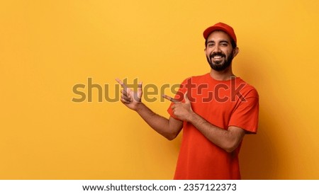 Happy Indian Courier Guy In Uniform Pointing Fingers Aside At Copy Space Handsome Smiling Eastern Man Advertising Delivery Service Offer, Standing Over Yellow Studio Background, Panorama Royalty-Free Stock Photo #2357122373