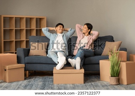 Apartment Rent Offer. Happy Korean Couple Resting Leaning On Couch In Modern Living Room At Home, Relaxing Among Packed Moving Boxes. Real Estate Property Purchase And Ownership Royalty-Free Stock Photo #2357122377