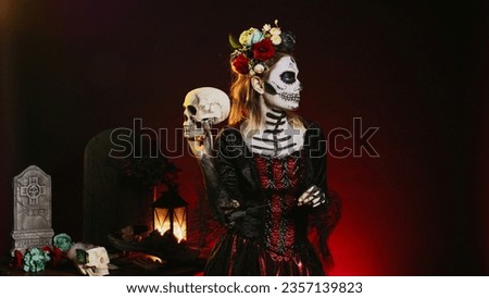 Horror scary model wearing traditional make up to celebrate mexican holiday, holding skull in studio. Looking like santa muerte or goddess of death on holy dios de los muertos celebration.