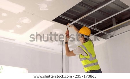 Asian construction worker in safety clothing and work gloves is fastening the drywall ceiling to the metal frame using an electric screwdriver on the ceiling covere Royalty-Free Stock Photo #2357173865