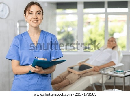 Positive young woman doctor posing against background of doctor's cabinet with patient lying on clinical chair Royalty-Free Stock Photo #2357178583