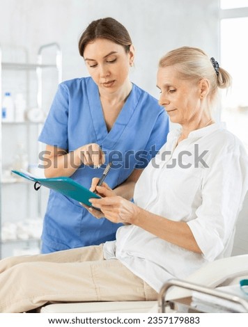 Old female patient lying on medical chair signing documents while talking with woman doctor Royalty-Free Stock Photo #2357179883