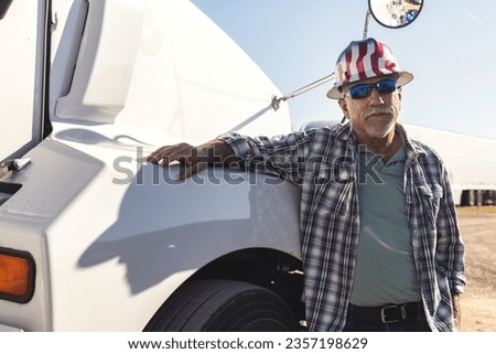 Portrait of old semi truck driver leaning on the hood of the semi-truck. Serious or worried trucker wearing safety helmet and sunglasses, next to his big rig. Royalty-Free Stock Photo #2357198629