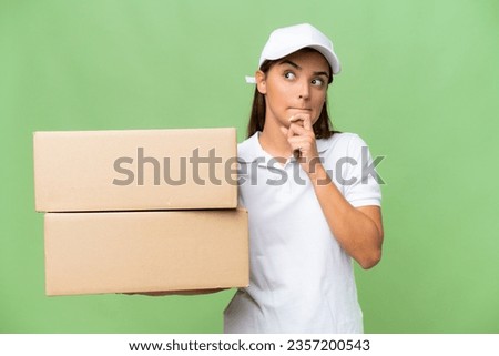Delivery caucasian woman holding boxes isolated on green chroma background having doubts and thinking