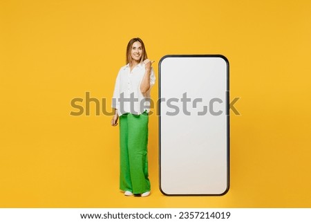 Full body young fun caucasian happy woman she wears white shirt casual clothes point htumb finger on big huge blank screen mobile cell phone smartphone with area isolated on plain yellow background