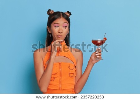 Asian woman with stylish hair buns and orange top holding glass of wine posing in blue studio, good mood concept, copy space