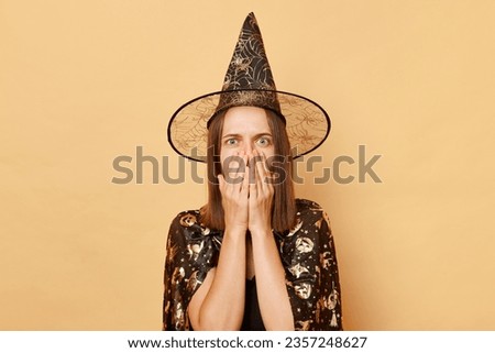 Shocked scared young girl wearing black halloween garment and cone posing isolated over beige background looking at camera with big eyes covering mouth scary holiday
