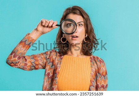 Investigator researcher scientist woman holding magnifying glass near face, looking into camera with big zoomed funny eyes, searching, analysing. Pretty young girl isolated on blue studio background Royalty-Free Stock Photo #2357251639
