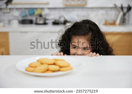 child girl hiding and looking butter cookies or biscuits on dish from under the table in the kitchen Royalty-Free Stock Photo #2357275219