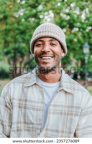 Vertical close up portrait of young hispanic man with a beanie hat smiling and looking at camera outdoors. Front view of latin happy guy standing at park with carefree attitude. Lifestyle concept Royalty-Free Stock Photo #2357276089