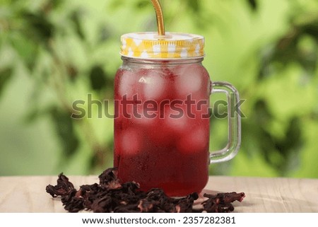 Refreshing hibiscus tea with ice cubes in mason jar and dry roselle flowers on wooden table against blurred green background