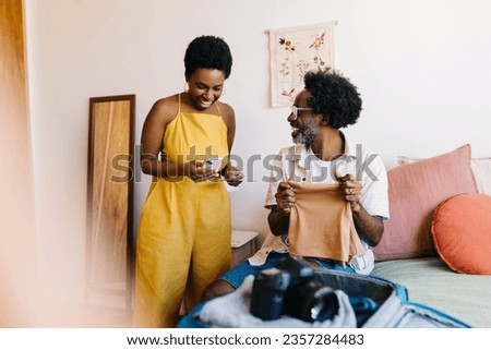 Happy couple in a bedroom, packing clothes for a romantic vacation and using a smartphone make travel arrangements. Mature couple planning and chatting, creating memories for their getaway together. Royalty-Free Stock Photo #2357284483
