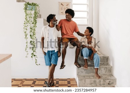 Teenage son and his parents, all with afro hair, laugh and smile together in a family moment. Happy black family relaxing by an indoor staircase, spending quality time and creating joyful memories. Royalty-Free Stock Photo #2357284957